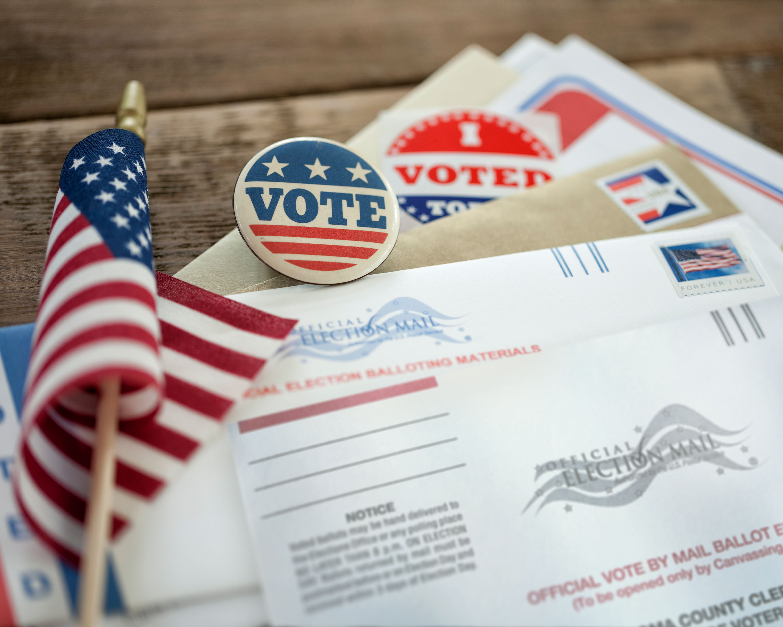 Cover image" photo of official election mail, a U.S. flag, and several "I Voted" stickers on a table. 